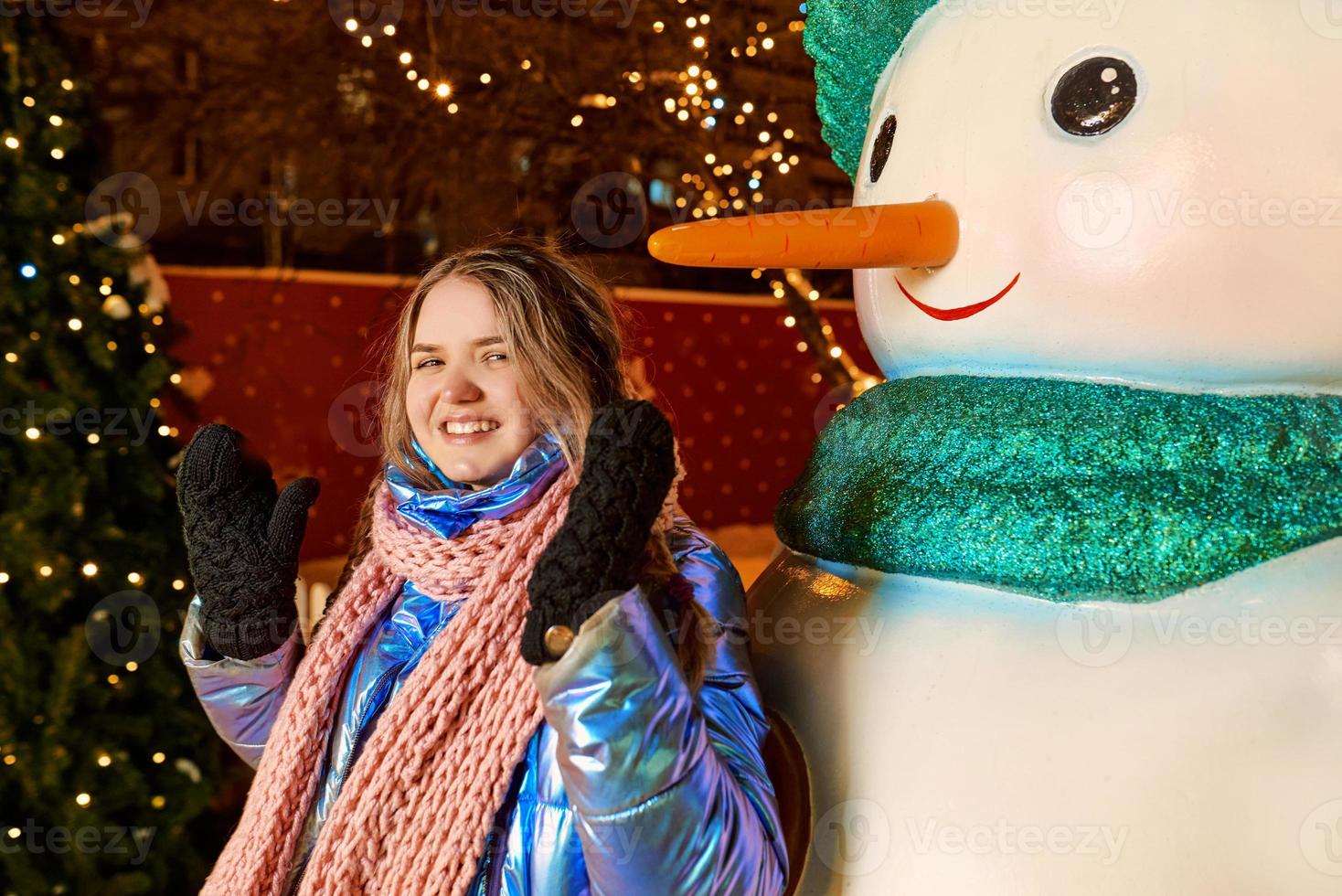 feliz sorridente jovem caucasiana no lenço, chapéu, jaqueta, luvas pelo boneco de neve ao ar livre. ano novo, diversão, conceito de inverno foto
