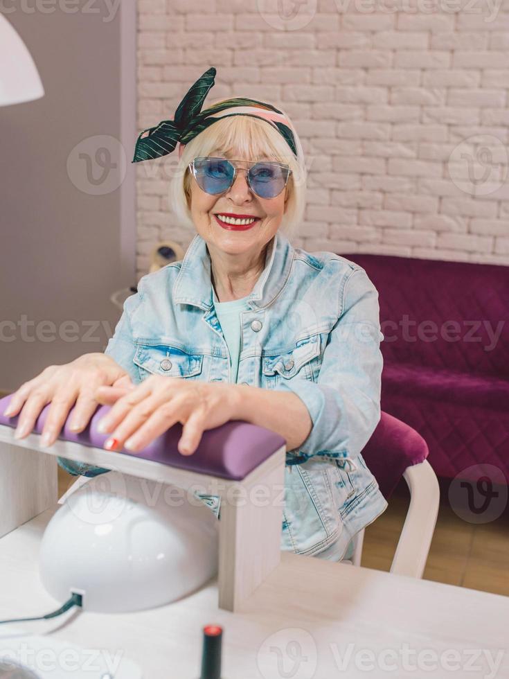 Mestre de manicure com luvas azuis cremosando as mãos de uma mulher idosa elegante em óculos de sol azuis e jaqueta jeans sentada no salão de manicure foto