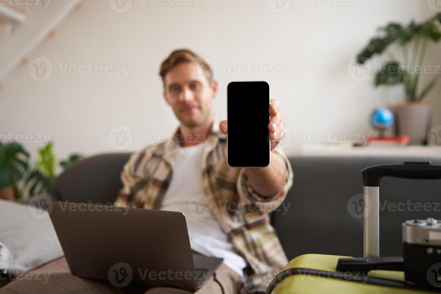 retrato do homem, turista mostra dele Móvel telefone tela, recomenda viagem aplicativo, sentado com mala de viagem e computador portátil foto