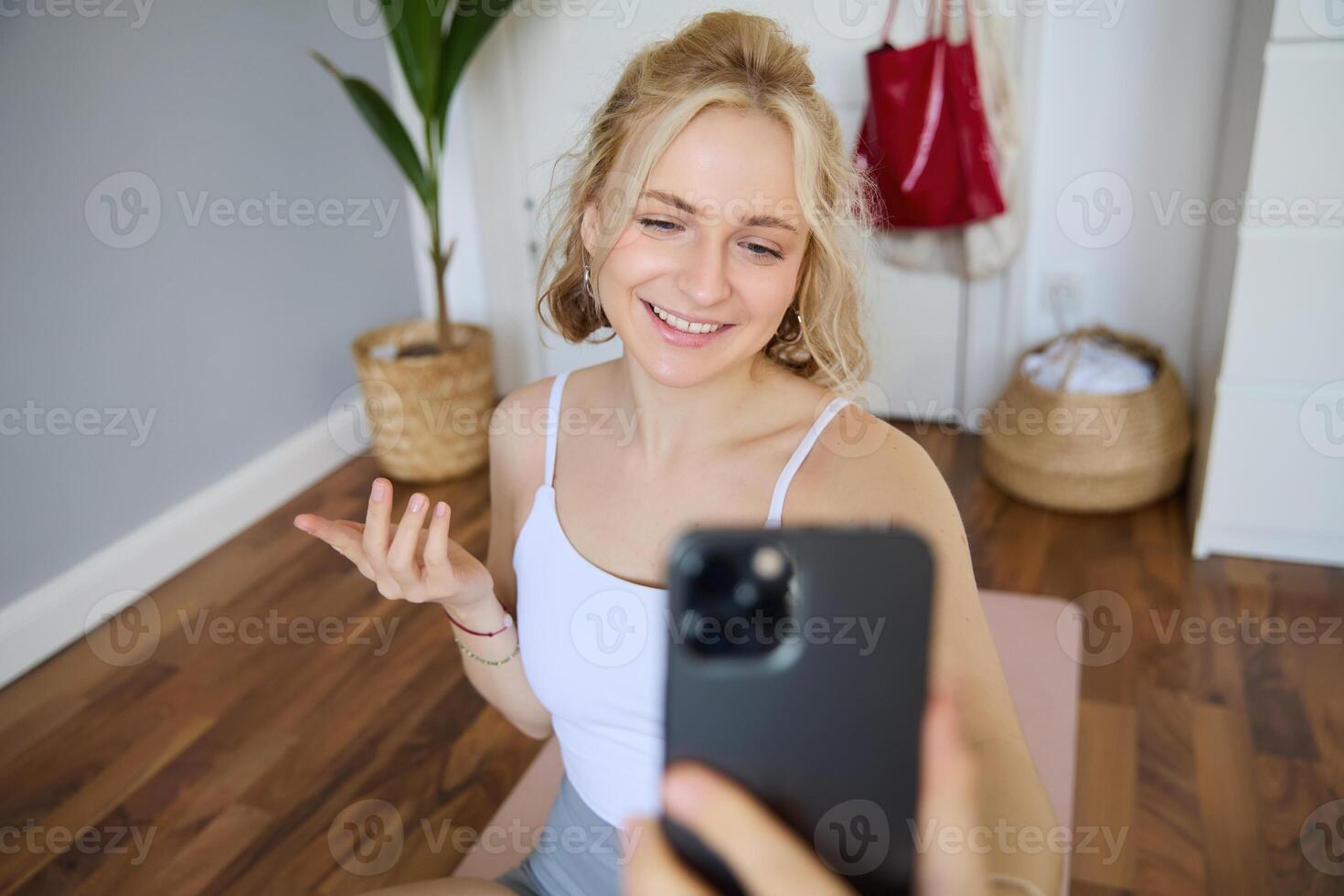 retrato do lindo jovem ginástica treinador, mostrando casa dar certo, levando selfie em Móvel telefone enquanto sentado em ioga borracha esteira dentro quarto, vestindo roupa ativa foto