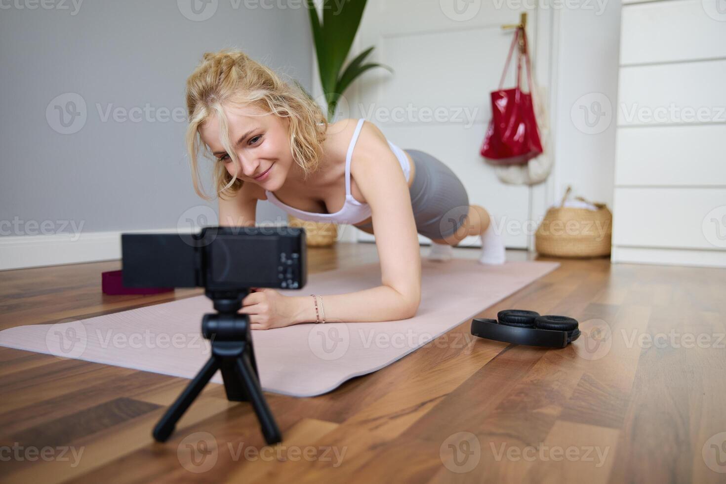jovem mulher, pessoal ginástica instrutor registros do ela mesma em pé dentro uma prancha, usando borracha ioga esteira e digital Câmera às casa foto