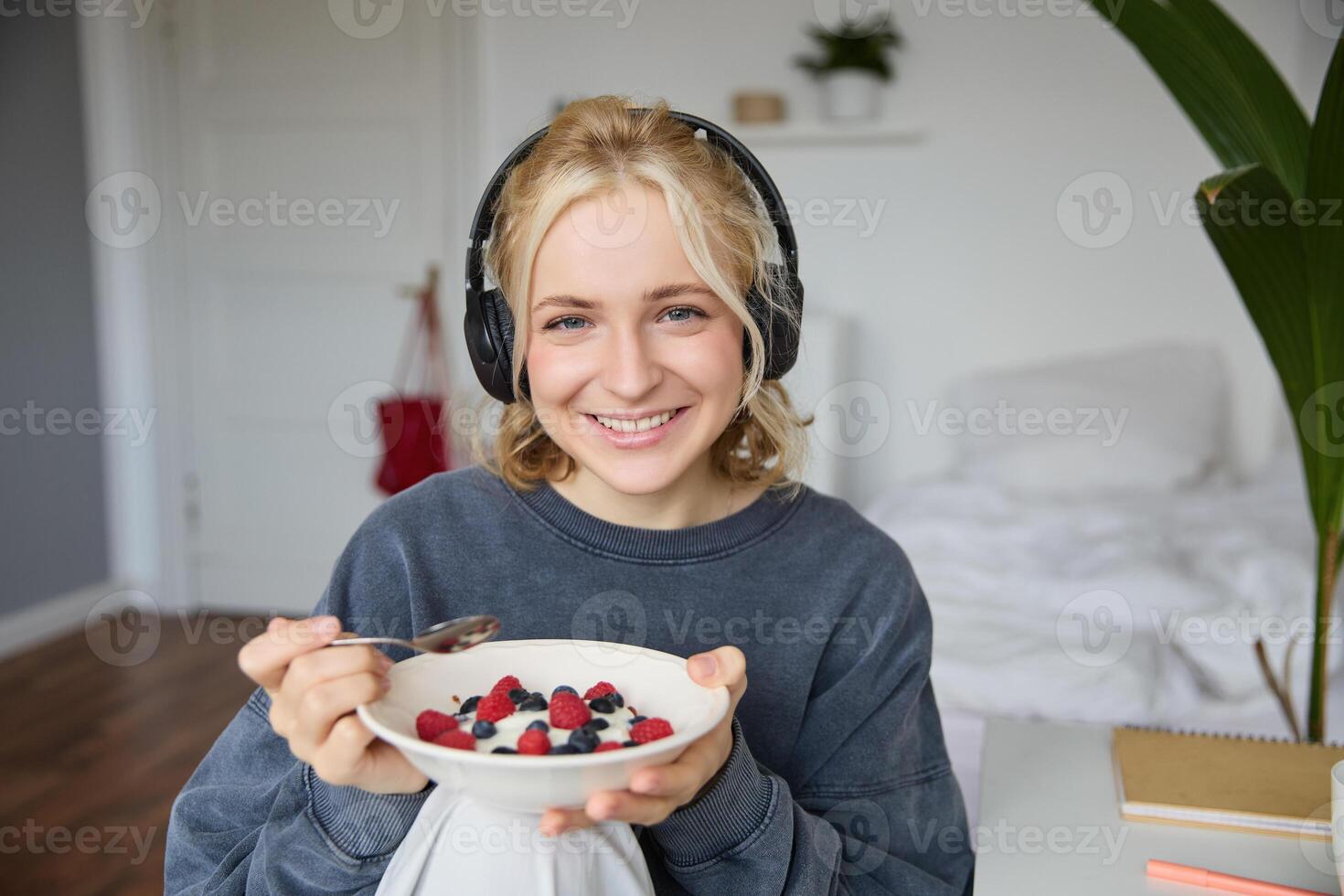 retrato do sorridente fofa mulher dentro fones de ouvido, comendo dela café da manhã e assistindo em computador portátil, olhando às tela com feliz face foto