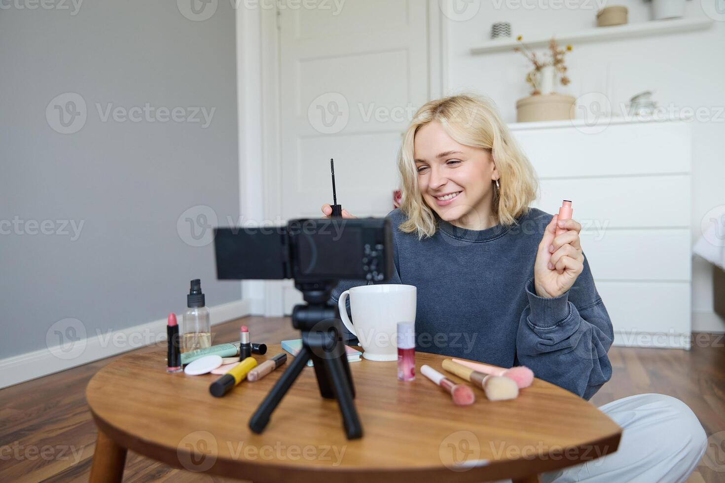 retrato do loiro sorridente mulher registros uma estilo de vida blog, vlogger ou Maquiagem artista gravação para social meios de comunicação, segurando rímel, revendo beleza produtos para seguidores conectados foto