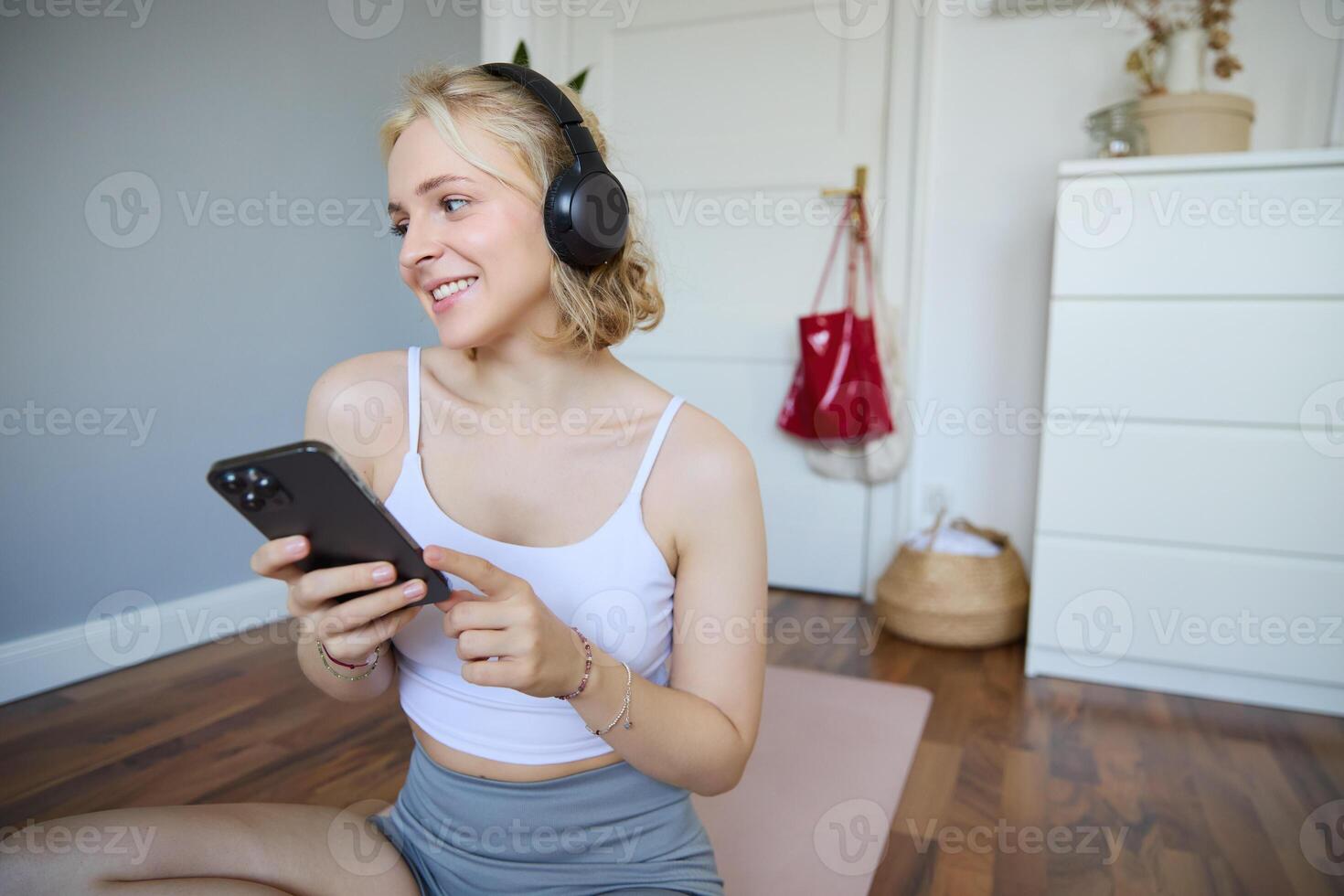 retrato do sorridente, saudável jovem mulher dentro fones de ouvido, segurando Smartphone, trabalhando Fora às lar, usando ginástica inscrição em Móvel telefone foto