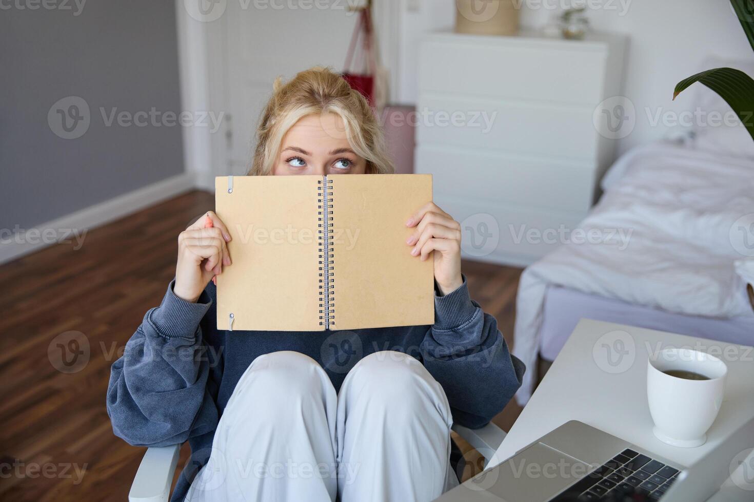 retrato do fofa loiro mulher, senta dentro frente do digital Câmera e computador portátil dentro dela sala, cobre face com caderno Diário, registros do ela mesma para estilo de vida vlog foto