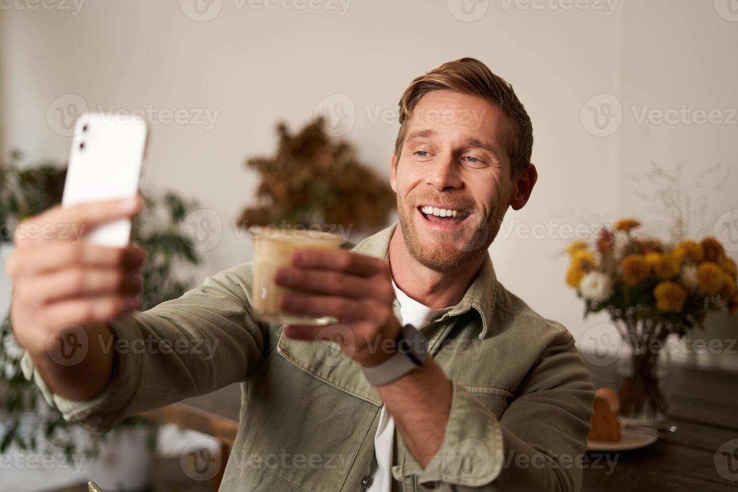 alegre bonito jovem homem, levando selfie em Móvel telefone aplicativo, chats para alguém e levantando vidro do café, Felicidades gesto, bebidas copo do cappuccino e sorrisos às Smartphone Câmera foto