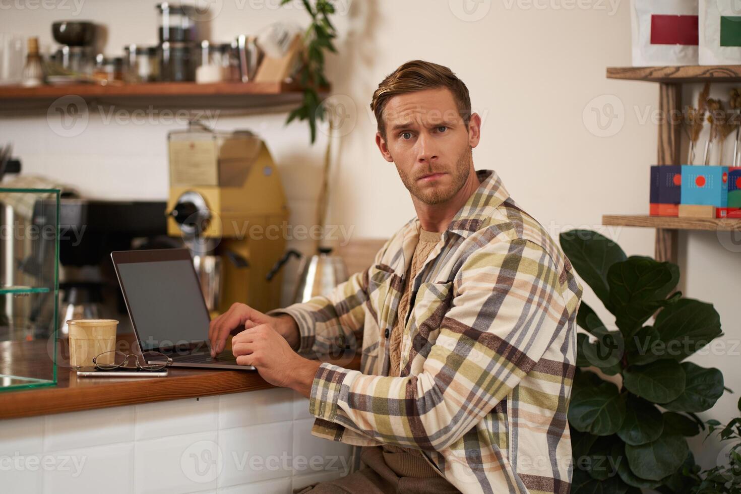 retrato do homem trabalhando dentro cafeteria, olhando confuso às Câmera, sentado com computador portátil e vidro do café, encarando intrigado às você foto