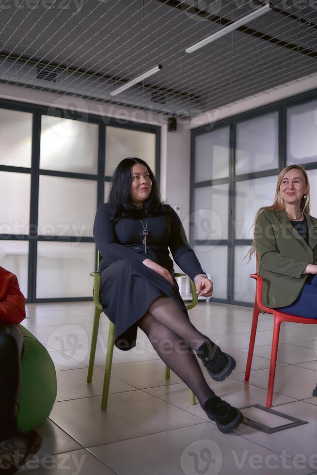 uma mulher faz uma discurso em estágio, colegas rir às dela piadas sentado em cadeiras e pufes dentro a salão, escritório localização foto