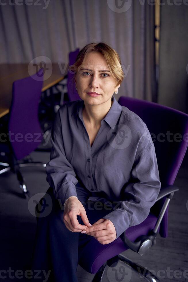 lindo meio era o negócio mulher vestindo cinzento camisa, Largo perna calça abd Preto estiletes sentar dentro roxa escritório cadeira dentro moderno área de trabalho foto