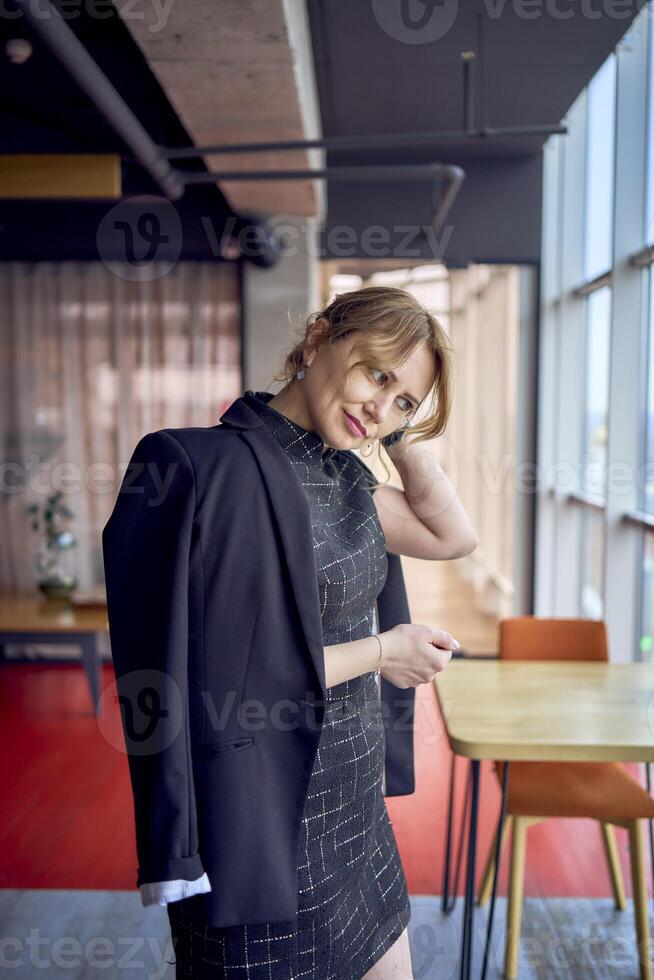 atraente mulher dentro uma Preto o negócio estilo mini vestir e Jaqueta vamos dela cabelo baixa dentro uma moderno loft estilo coworking espaço foto