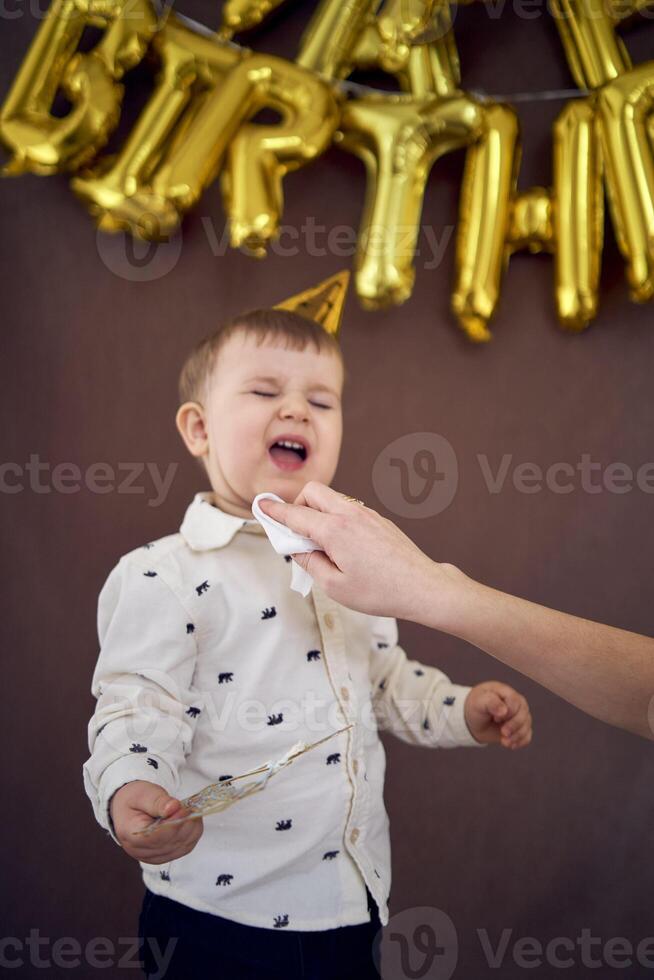 uma 2 anos de idade Garoto é espalhafatoso quando dele mãe tentativas para limpar ele fora a aniversário bolo foto