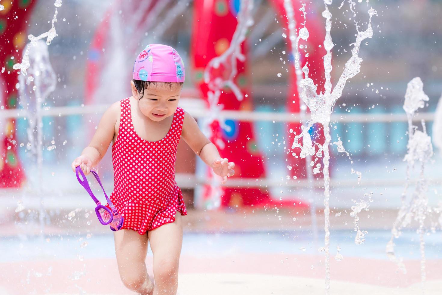 criança feliz brincar com água para esfriar dançando com fonte no parque aquático no verão ou primavera. garota se diverte jogando em fontes de água. criança usa maiô vermelho. bebê de 3 anos. foto