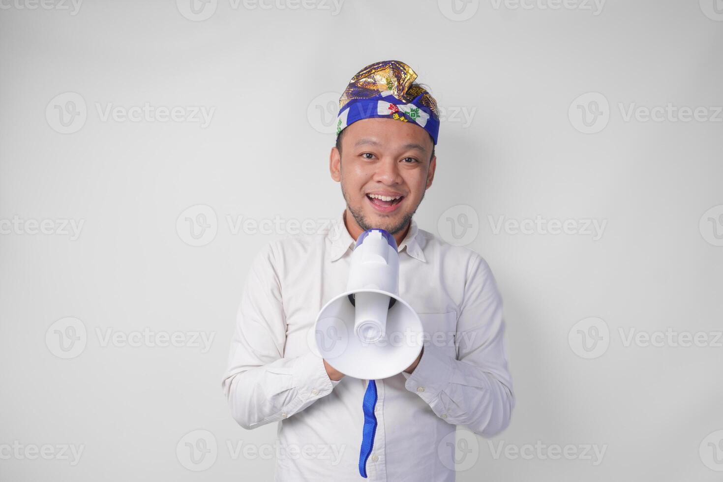 enérgico balinesa homem dentro branco camisa e tradicional cocar gritando às megafone enquanto levantando cerrado punho, isolado de branco fundo foto