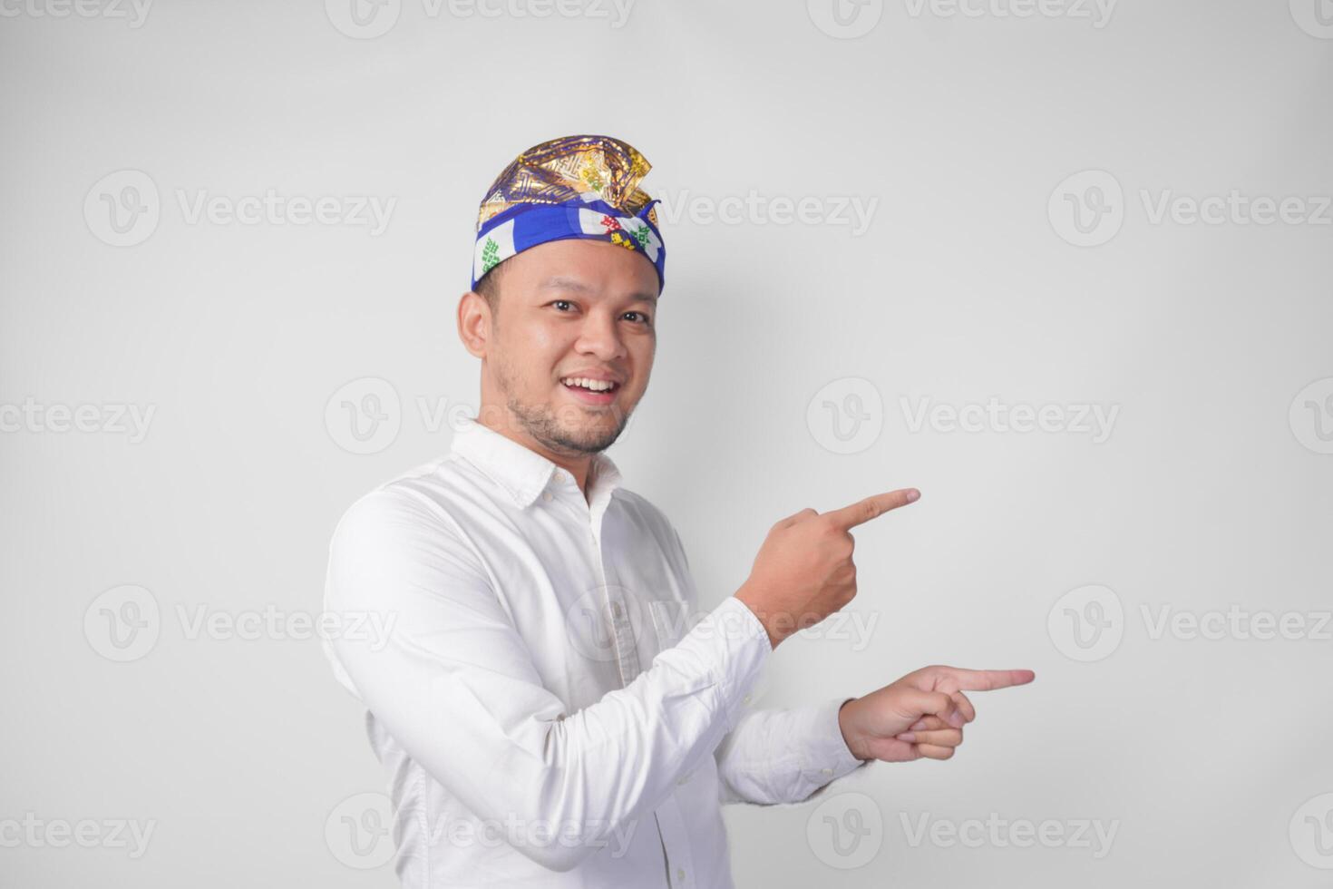 atraente balinesa homem dentro branco camisa e tradicional cocar chamado udeng apontando para a cópia de espaço em a esquerda e certo lado enquanto sorridente alegremente foto