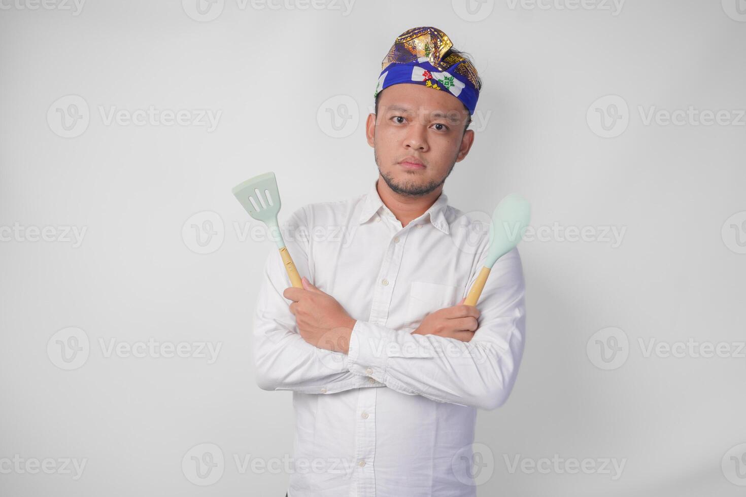 jovem balinesa homem dentro branco camisa e tradicional cocar segurando espátula e cozinha cozinhando utensílios com sério expressão foto