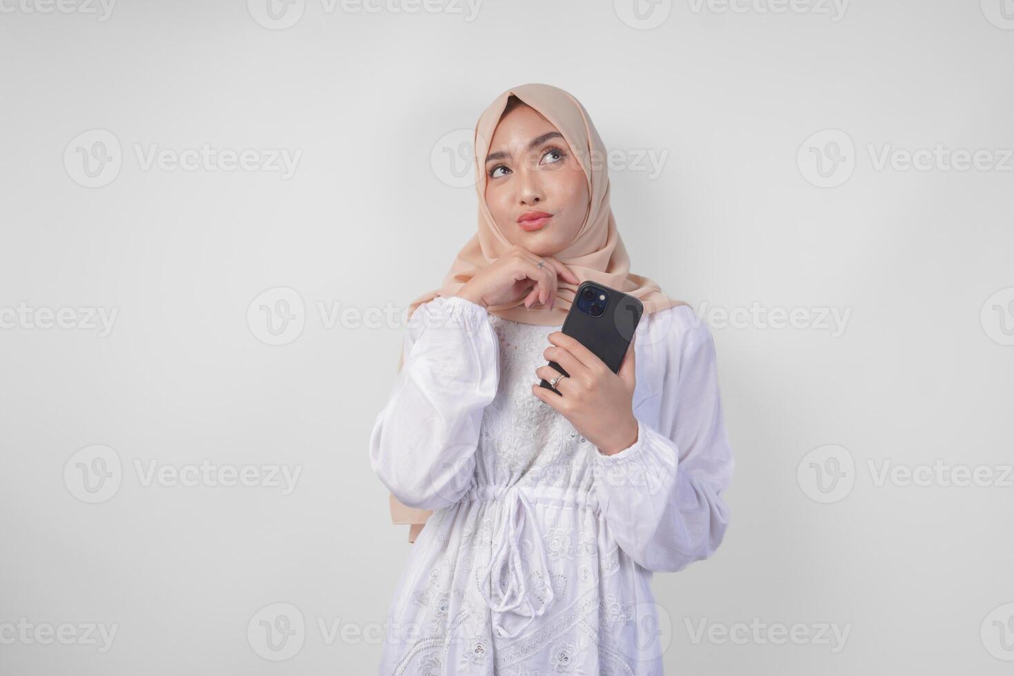 pensativo jovem ásia muçulmano mulher vestindo branco vestir e hijab, usando Smartphone enquanto segurando dela queixo e pensando com sério expressão sobre isolado branco fundo foto