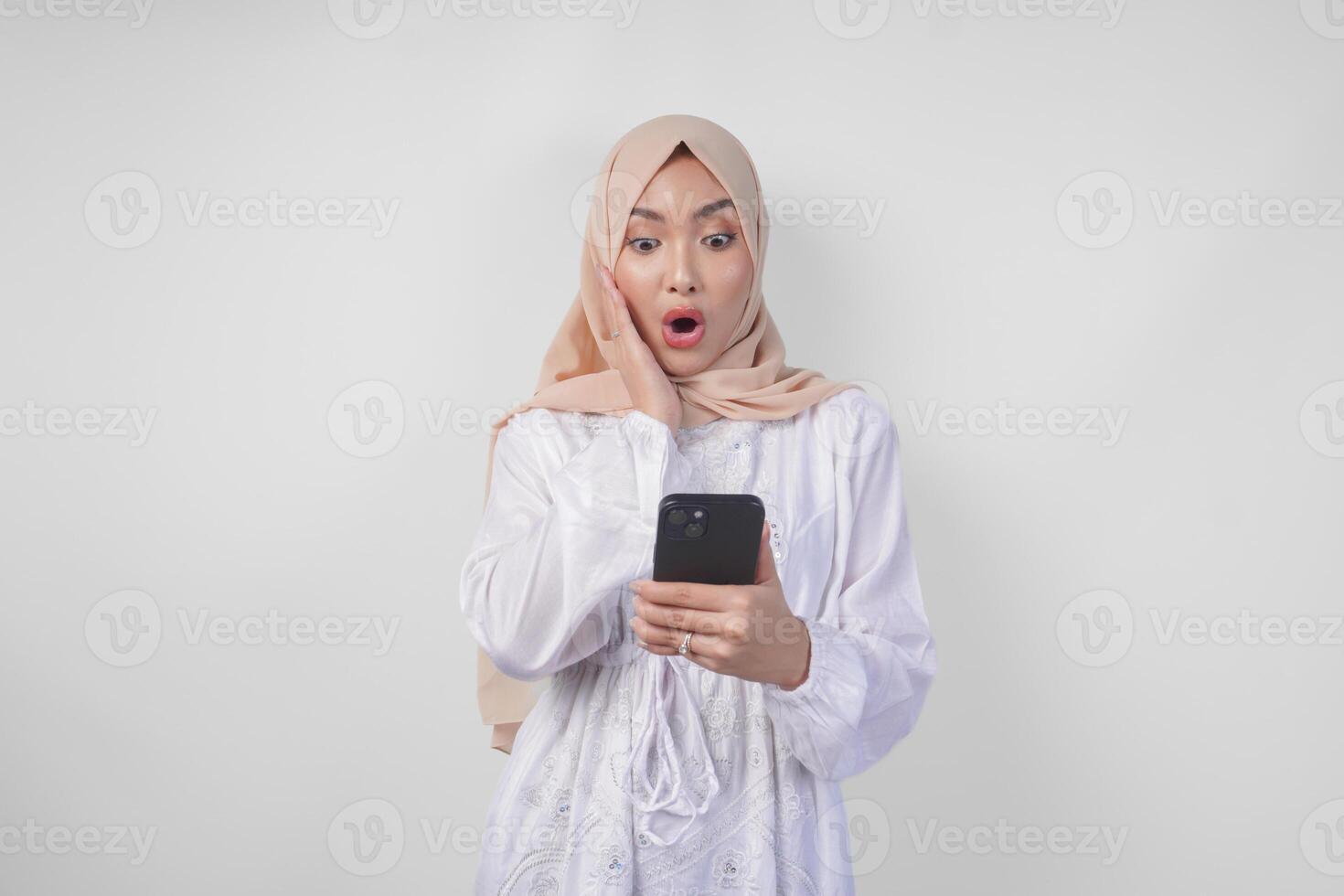 jovem ásia muçulmano mulher vestindo branco vestir e hijab mostrando chocado expressão em dela face enquanto segurando Smartphone, surpreso depois de lendo notícia ou fofoca, isolado de branco fundo foto