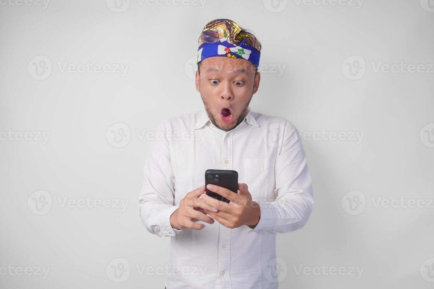 jovem balinesa homem vestindo branco camisa e tradicional cocar mostrando chocado expressão em face enquanto segurando Smartphone, surpreso depois de lendo notícia ou fofoca, isolado de branco fundo foto