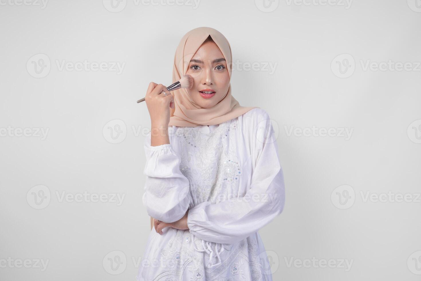 lindo jovem ásia muçulmano mulher vestindo branco vestir e hijab aplicando faço acima usando escovar. moda e cosméticos beleza conceito foto