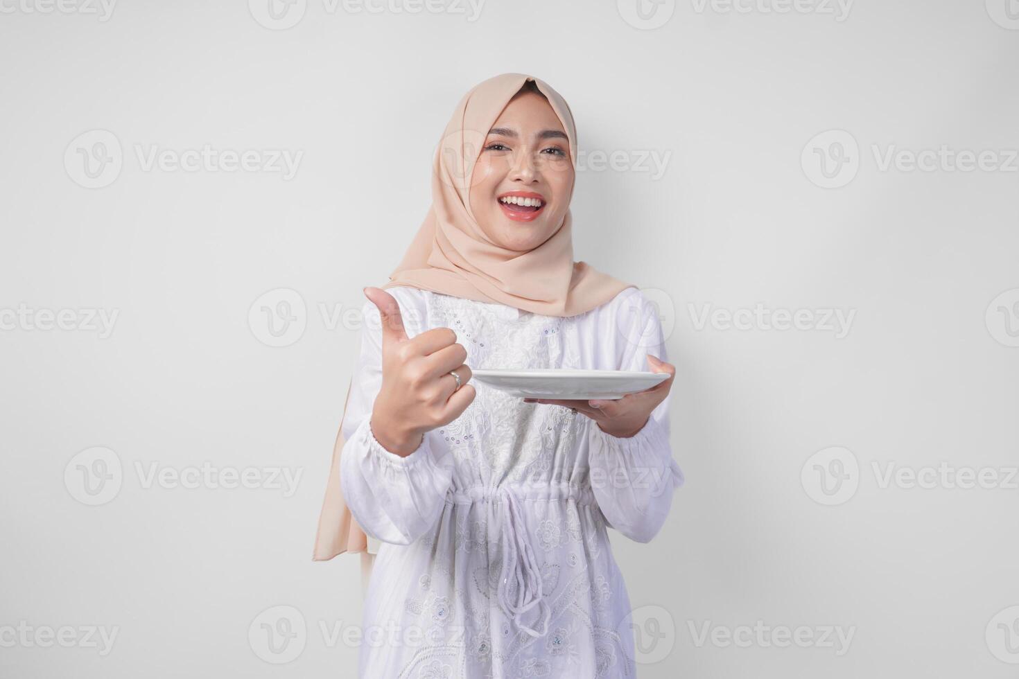 animado jovem ásia muçulmano mulher dentro hijab apresentando esvaziar branco prato cópia de espaço enquanto fazendo polegares acima gesto foto