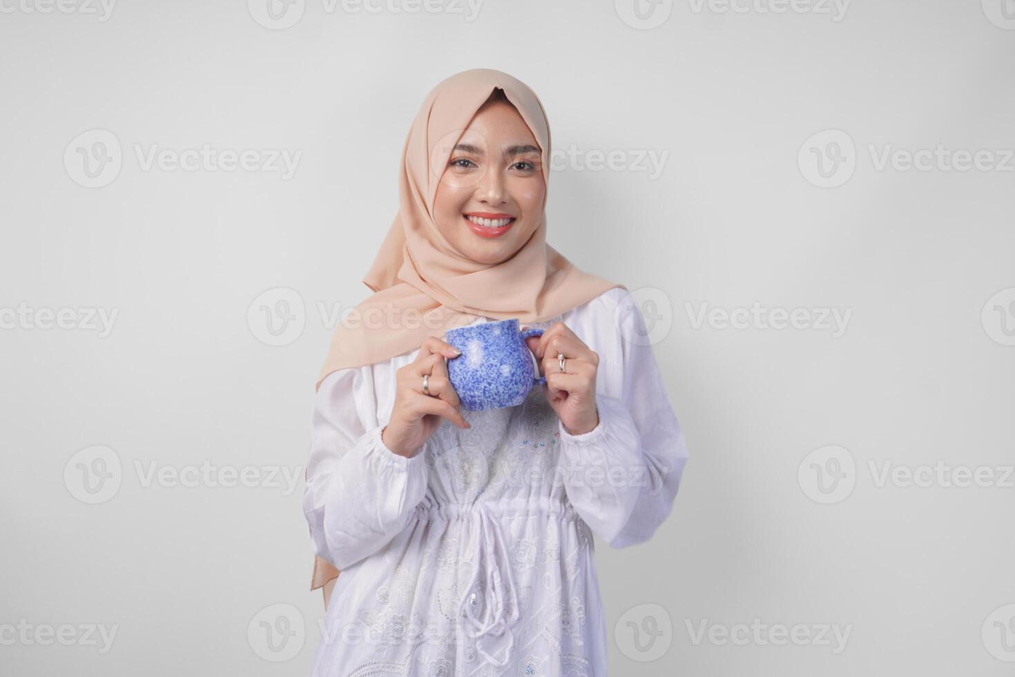 sorridente ásia muçulmano mulher dentro branco vestir e hijab segurando e apresentando água ou bebida dentro uma azul xícara, em pé sobre isolado branco fundo foto