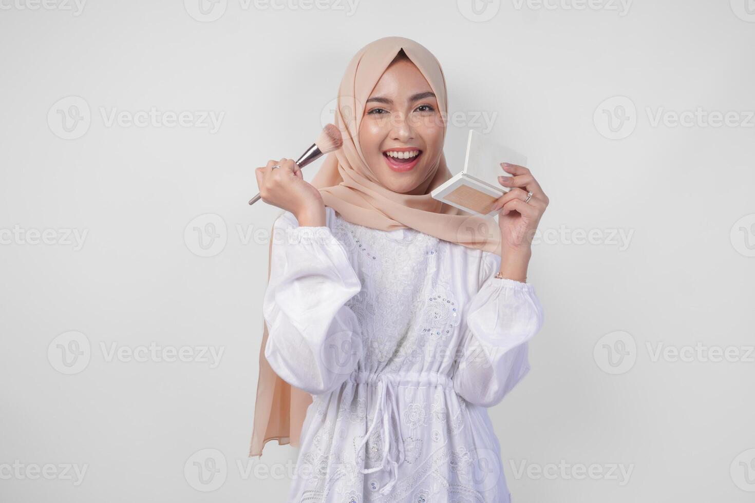 lindo jovem ásia muçulmano mulher vestindo branco vestir e hijab aplicando faço acima usando escovar. moda e cosméticos beleza conceito foto