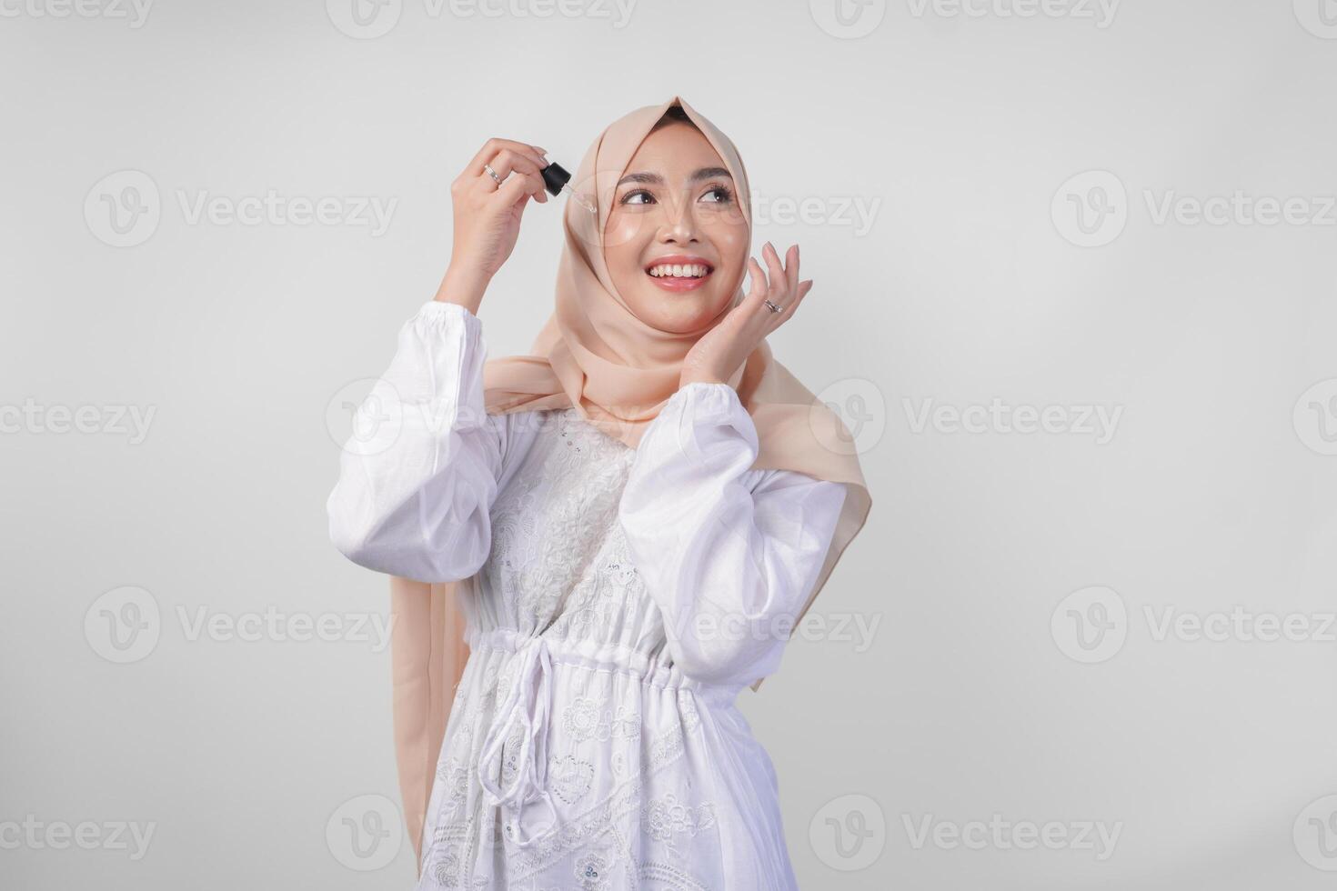 animado jovem ásia muçulmano mulher vestindo branco vestir e hijab teste ou aplicando pele Cuidado sérum em face. facial e beleza conceito foto