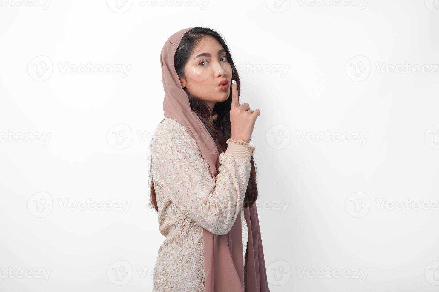 jovem ásia muçulmano mulher vestindo lenço de cabeça véu hijab olhando para Câmera enquanto colocando uma dedo dentro frente do lábios gesticulando fique silencioso ou fique quieto, isolado de branco fundo foto