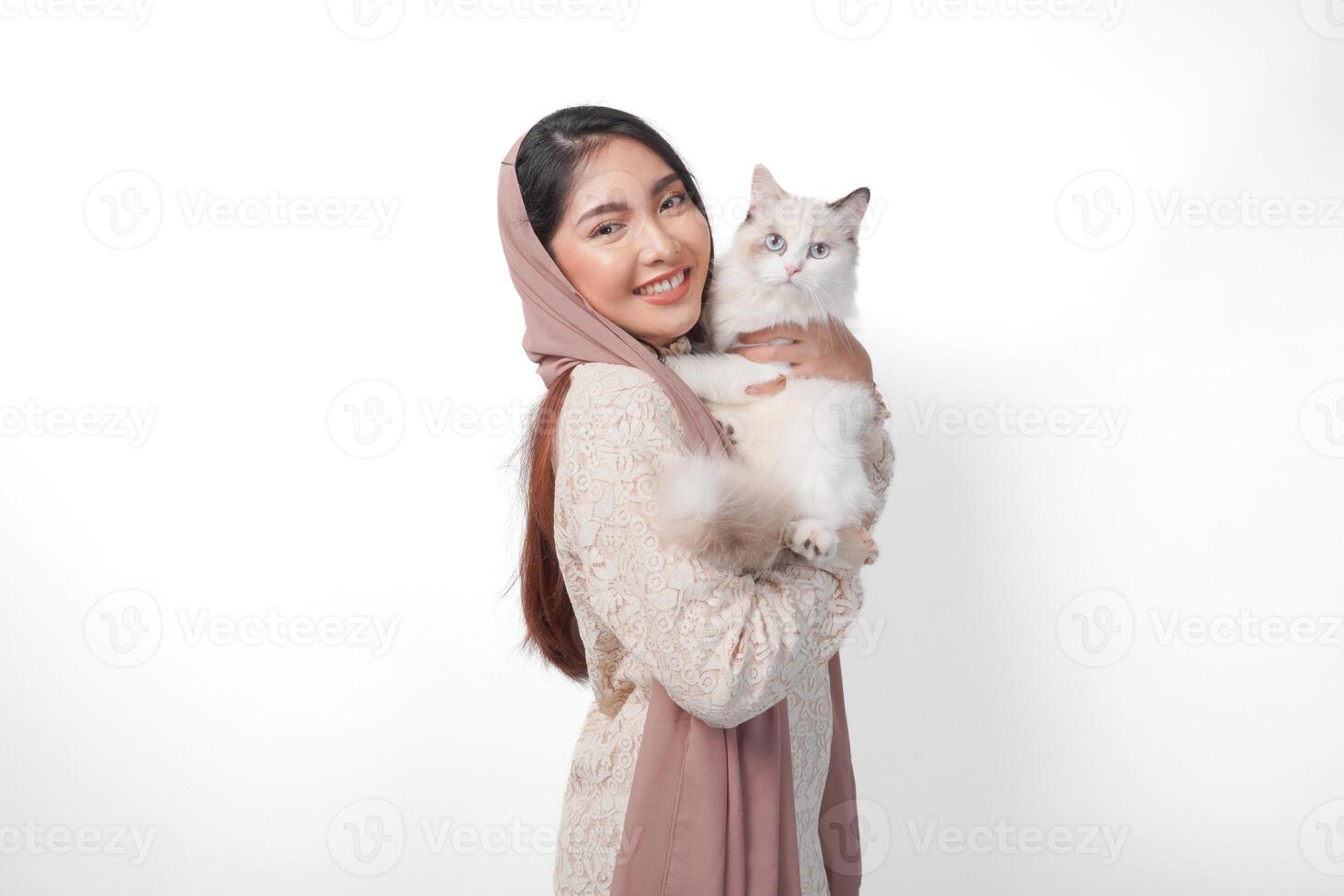 atraente jovem ásia muçulmano mulher dentro véu hijab sorridente enquanto posando e abraçando uma branco boneca de pano gato animal foto