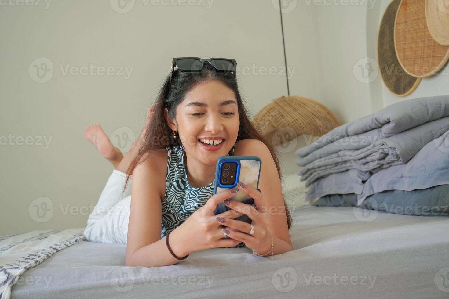 sorridente ásia mulher deitado baixa em cama com oculos de sol em topo enquanto usando Móvel telefone, explorando social meios de comunicação dentro a manhã foto