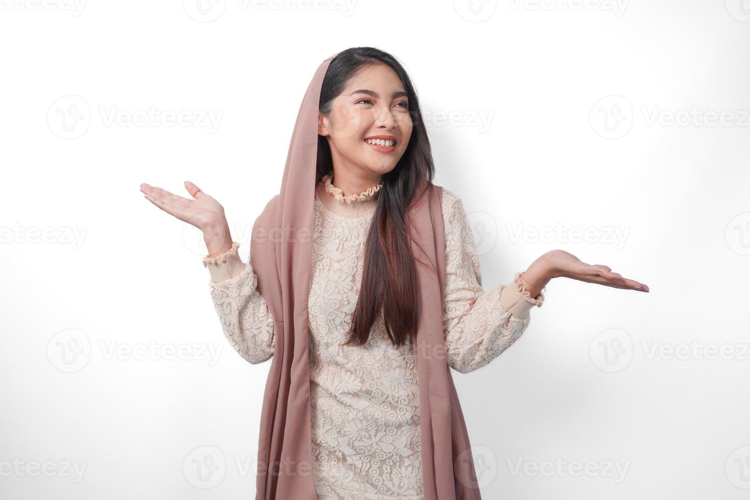 ásia muçulmano mulher vestindo lenço de cabeça véu hijab sorridente alegremente enquanto olhando e apontando para cópia de espaço ao lado dela, em pé em isolado branco fundo foto
