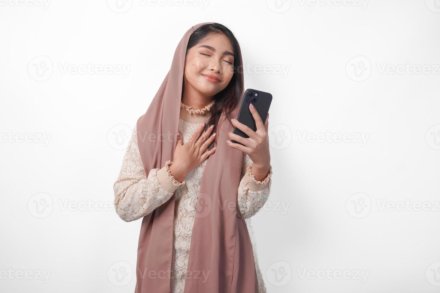 feliz atento grato jovem ásia muçulmano mulher mão em peito sorridente enquanto segurando Smartphone, isolado em branco fundo foto