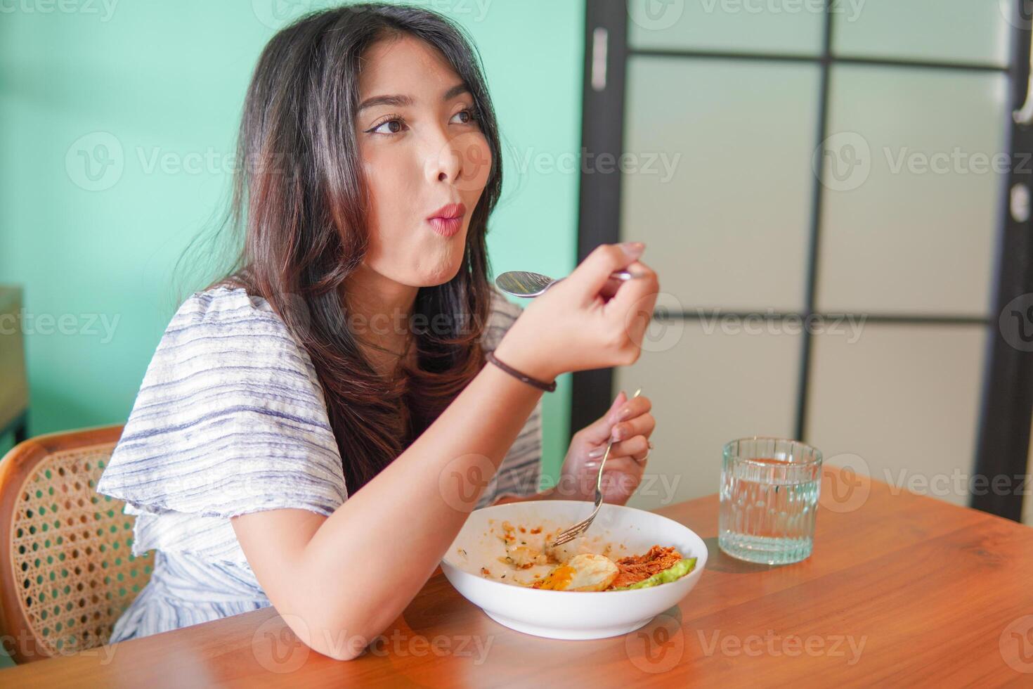 retrato do a animado jovem ásia mulher vestindo vestir sentado às uma restaurante, comendo e desfrutando café da manhã com uma alegre sorrir foto