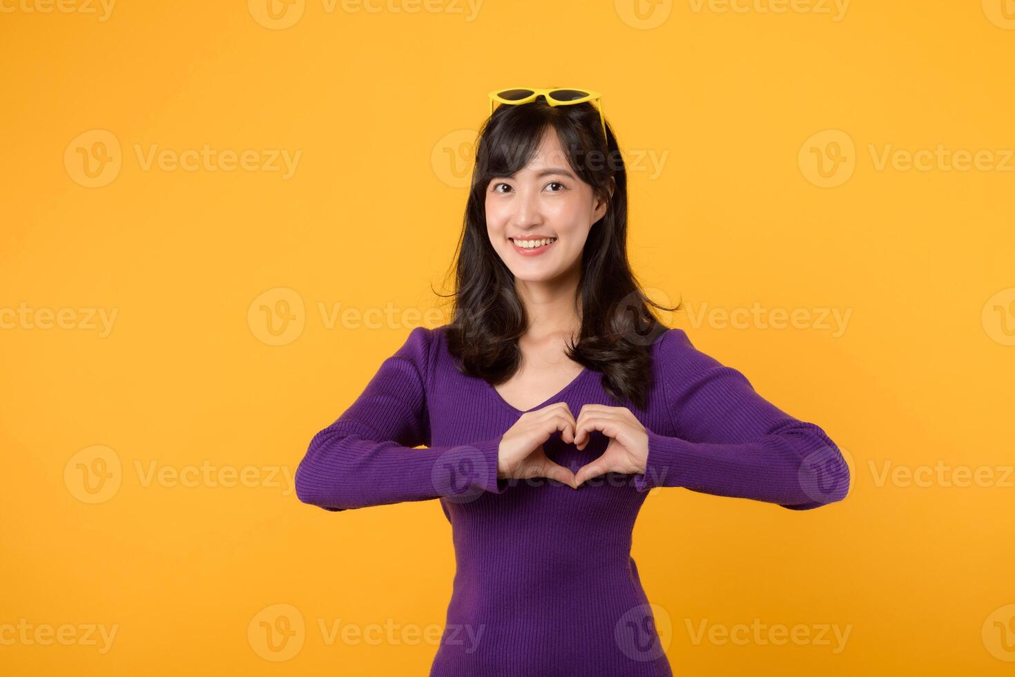 contra uma amarelo fundo, expressando amor e felicidade, uma alegre jovem mulher, vestindo uma roxa camisa e Óculos, formulários uma coração forma com dela mão dentro uma estúdio retrato, simbolizando romance. foto