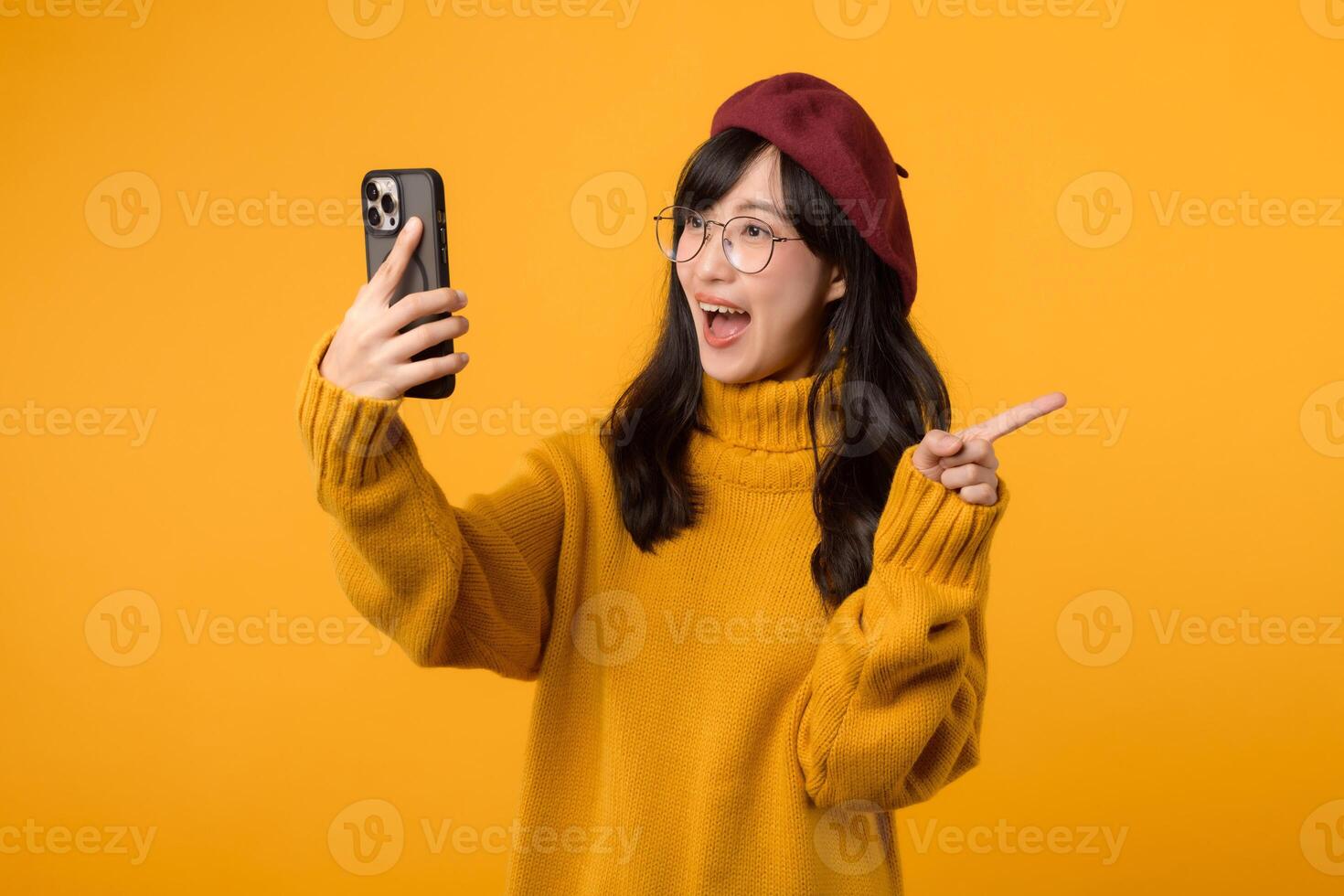 retrato jovem lindo 30s ásia mulher feliz sorrir vestindo amarelo camisa e vermelho chapéu apontando dedo gesto para livre cópia de espaço e usando Smartphone contra amarelo fundo. Novo inscrição foto