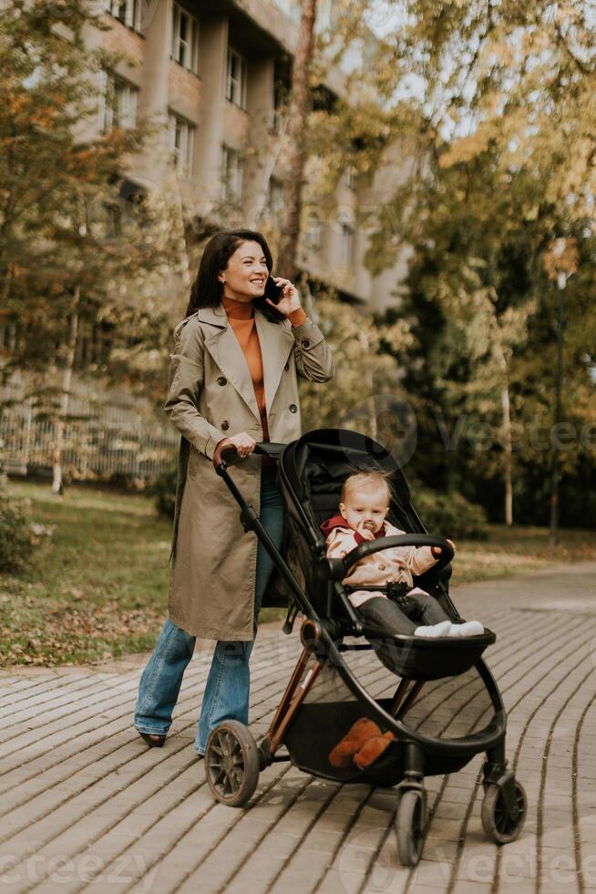 jovem mulher com fofa bebê menina dentro bebê carrinho de criança usando Móvel telefone às a outono parque foto