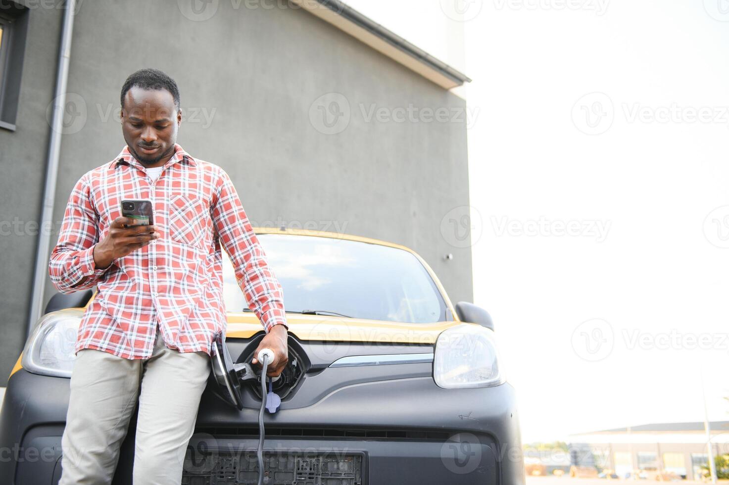 casual africano homem com Smartphone perto elétrico carro esperando para a terminar do a bateria cobrando processo. foto