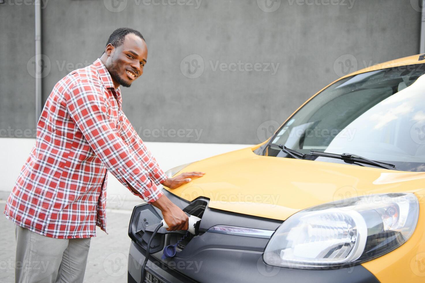 uma africano americano homem carrinhos Próximo para amarelo elétrico Entrega furgão às elétrico veículo cobrando estações foto