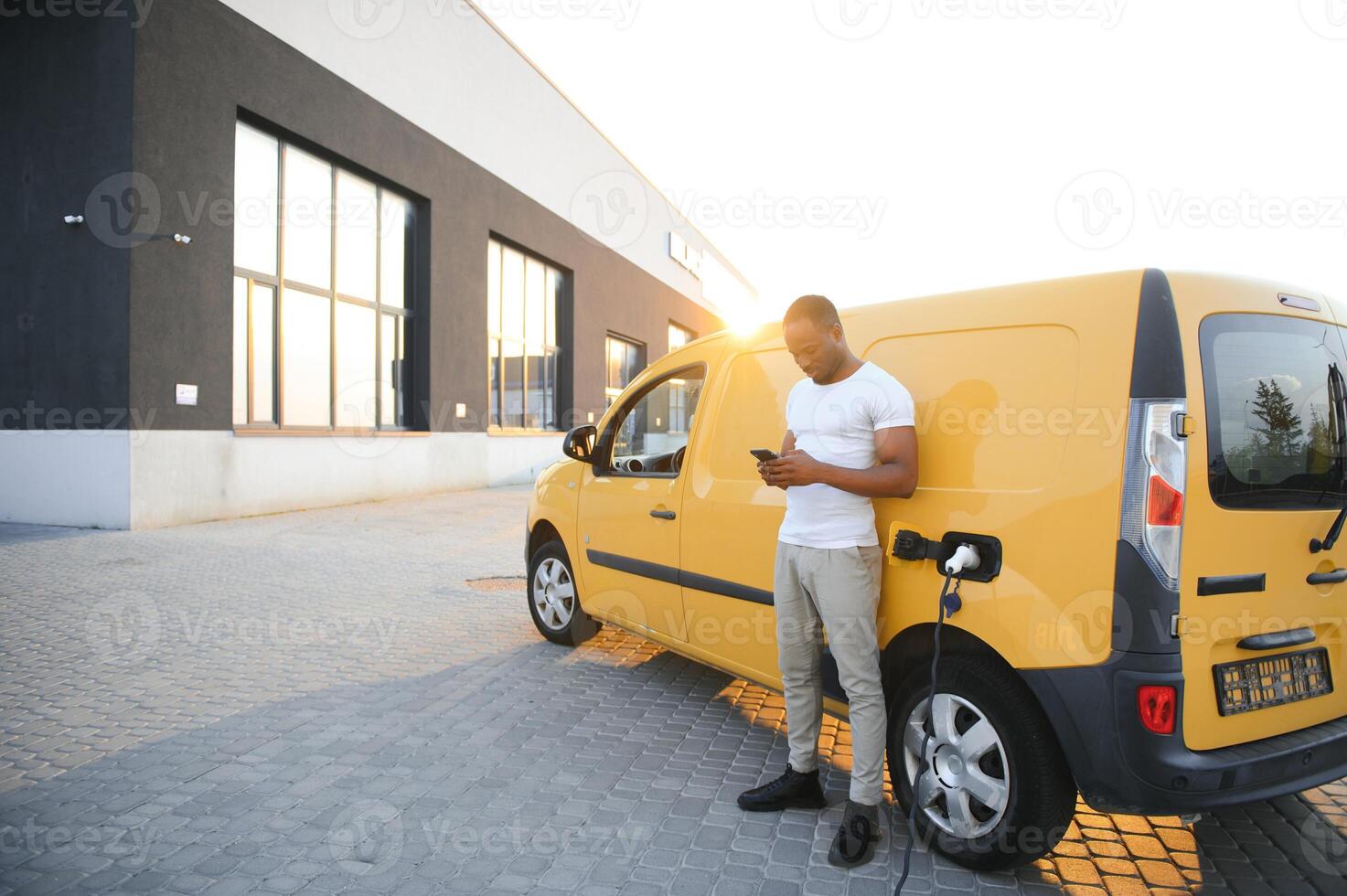 casual africano homem com Smartphone perto elétrico carro esperando para a terminar do a bateria cobrando processo. foto
