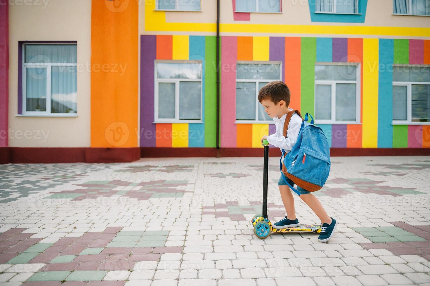 Adolescência Garoto com pontapé lambreta perto moderno escola. criança com mochila e livro ao ar livre. começando do aulas. primeiro dia do cair. costas para escola. foto