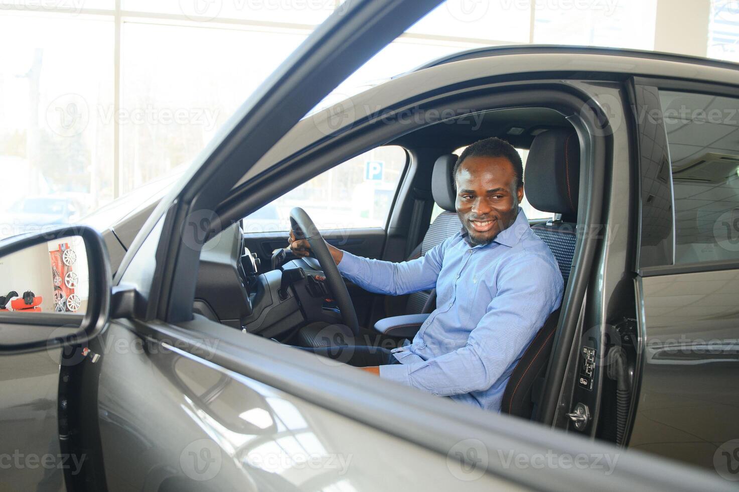 feliz carro comprador, Novo carro proprietário conceito. retrato do animado jovem africano americano cara dentro concessionária sala de exposições foto