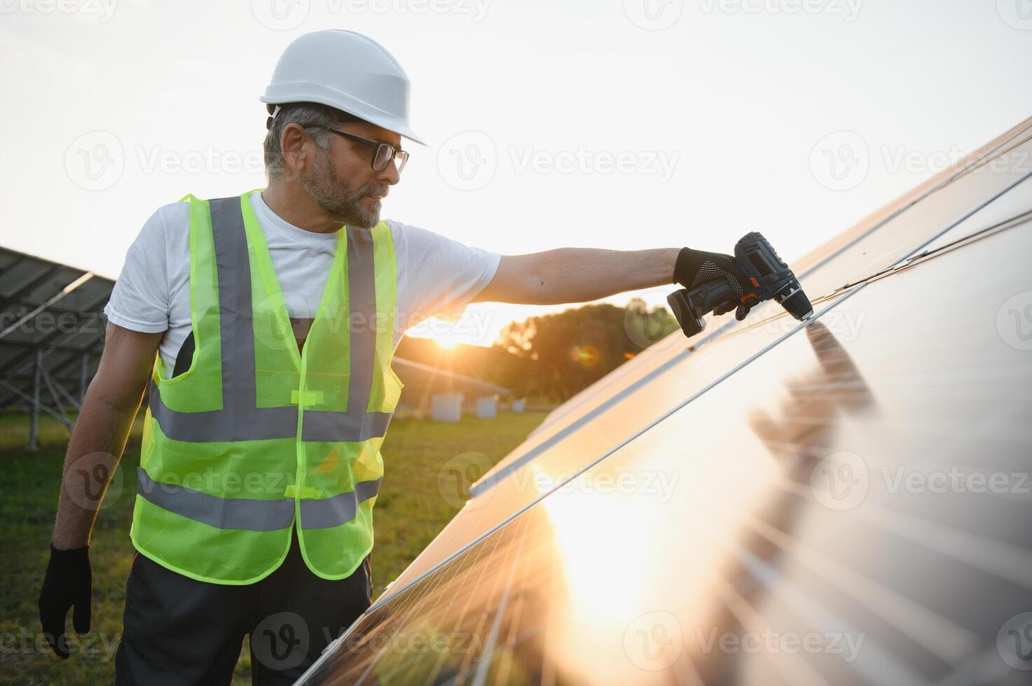 retrato do sorridente confiante engenheiro técnico com elétrico Chave de fenda, em pé dentro frente do inacabado Alto exterior solar painel foto voltaico sistema