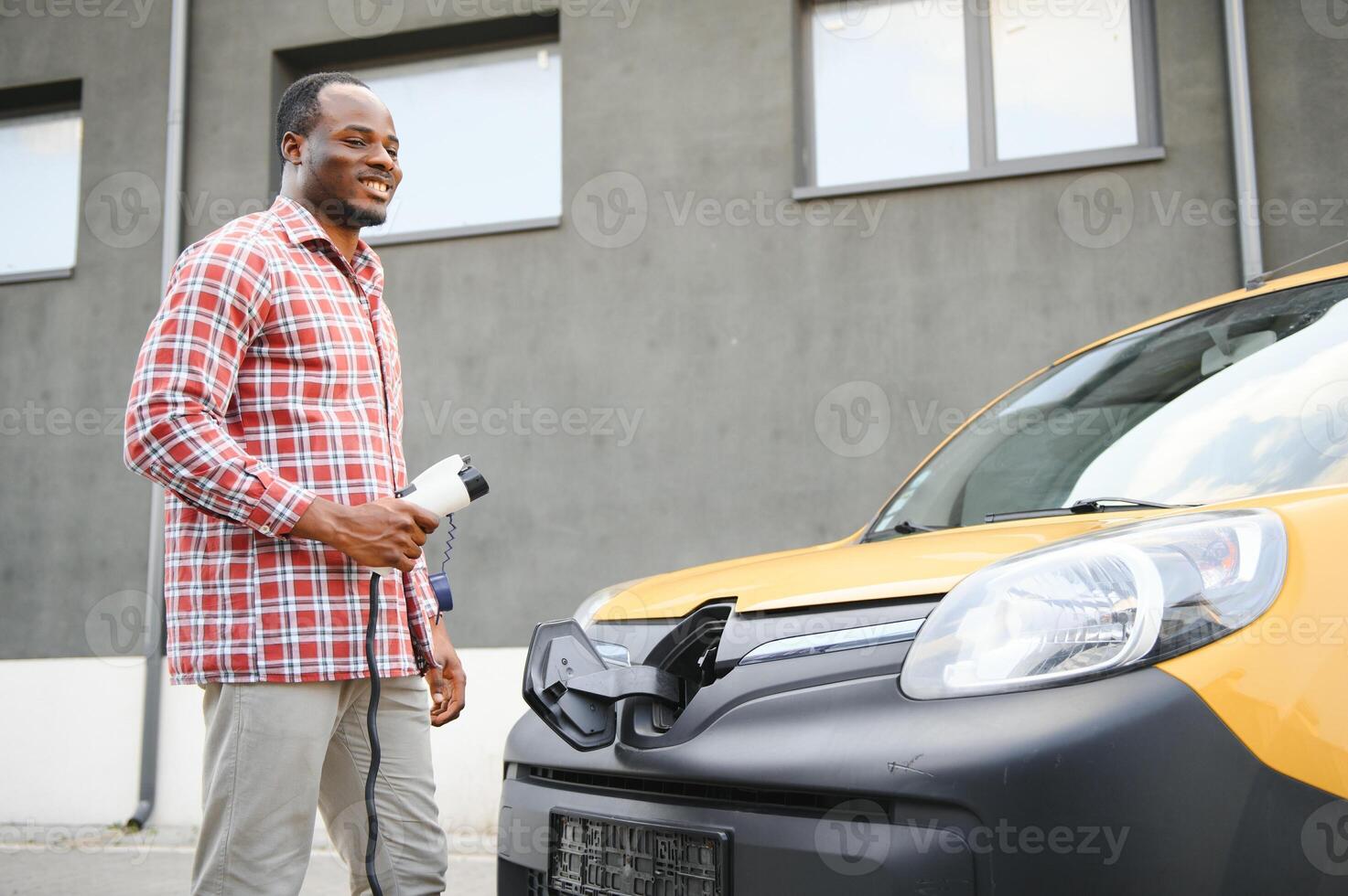 uma africano americano homem carrinhos Próximo para amarelo elétrico Entrega furgão às elétrico veículo cobrando estações foto