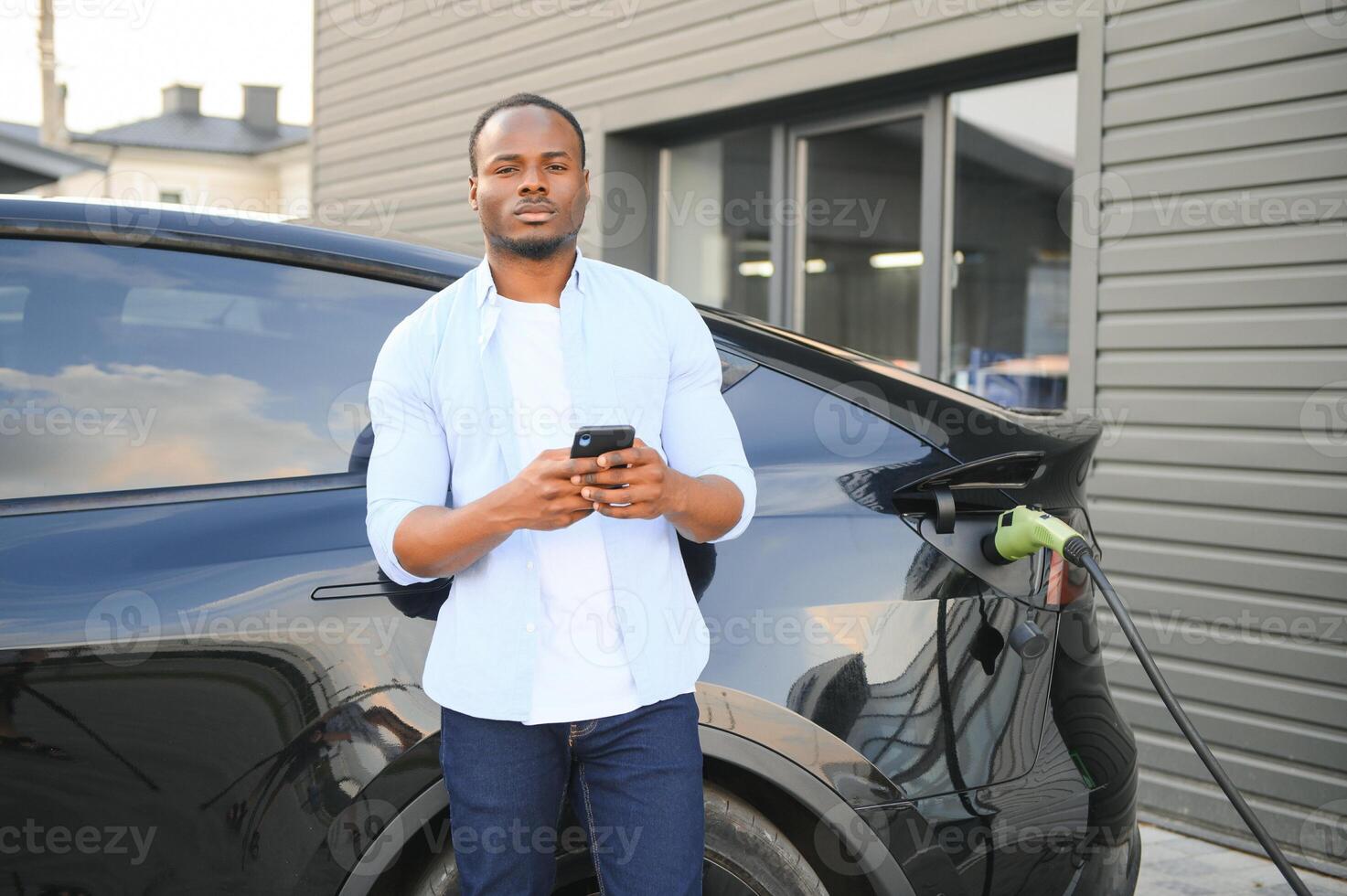 a africano homem é em pé perto a elétrico carro, esperando para isto para carregar às uma cobrando estação e usando a telefone. foto