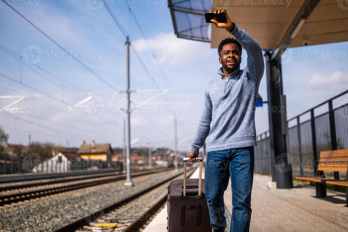 homem acenando para uma deixando trem e corrida ao longo estrada de ferro estação com mala de viagem e Móvel telefone. foto