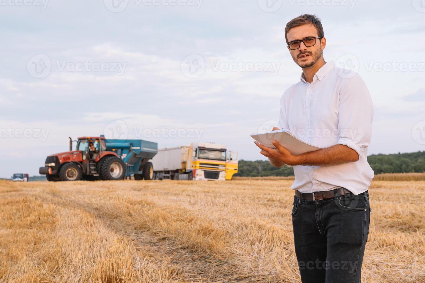 jovem atraente agricultor com computador portátil em pé dentro trigo campo com combinar colheitadeira dentro fundo. foto