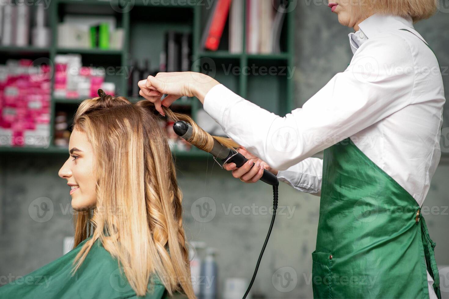 criando cachos com ondulação ferros. cabeleireiro faz uma Penteado para uma jovem mulher com grandes vermelho cabelo dentro uma beleza salão. foto