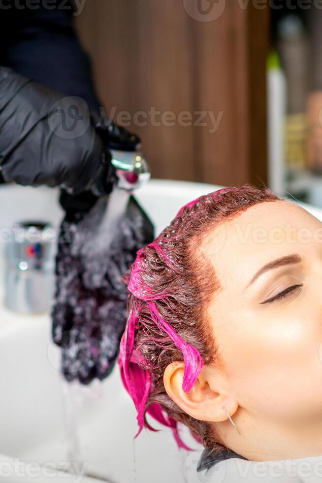 lavando tingido fêmea cabelo. profissional cabeleireiro lavagens Rosa cor pintura fora do uma cliente. jovem caucasiano mulher tendo dela cabelo lavado dentro uma beleza salão. foto