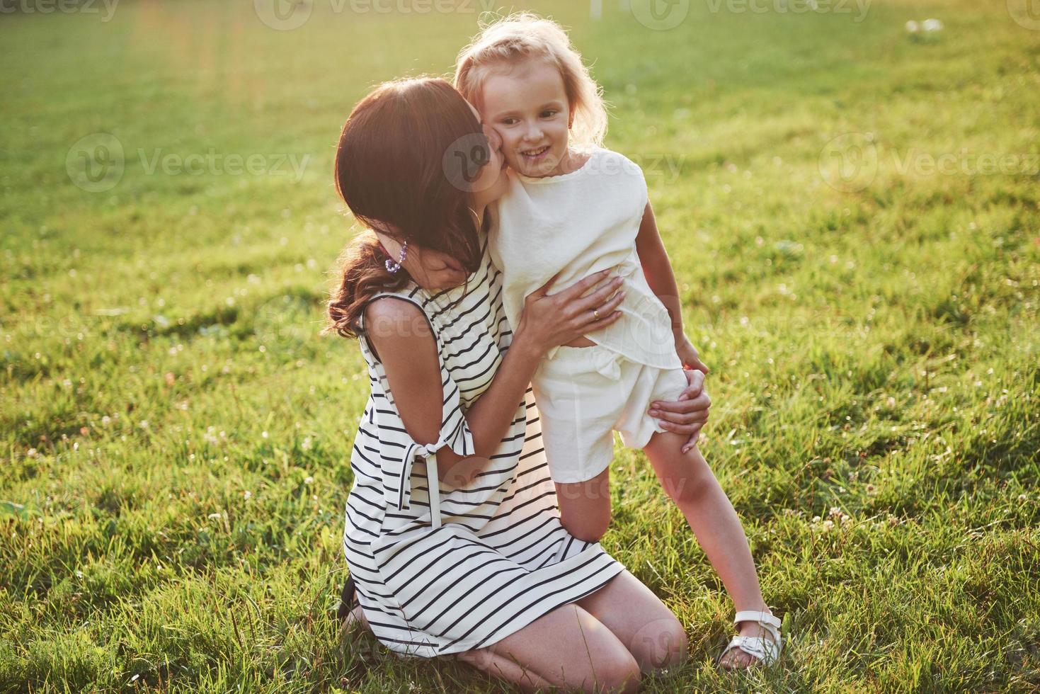 feliz mãe e filha se abraçando em um parque ao sol em um fundo de verão brilhante de ervas. foto