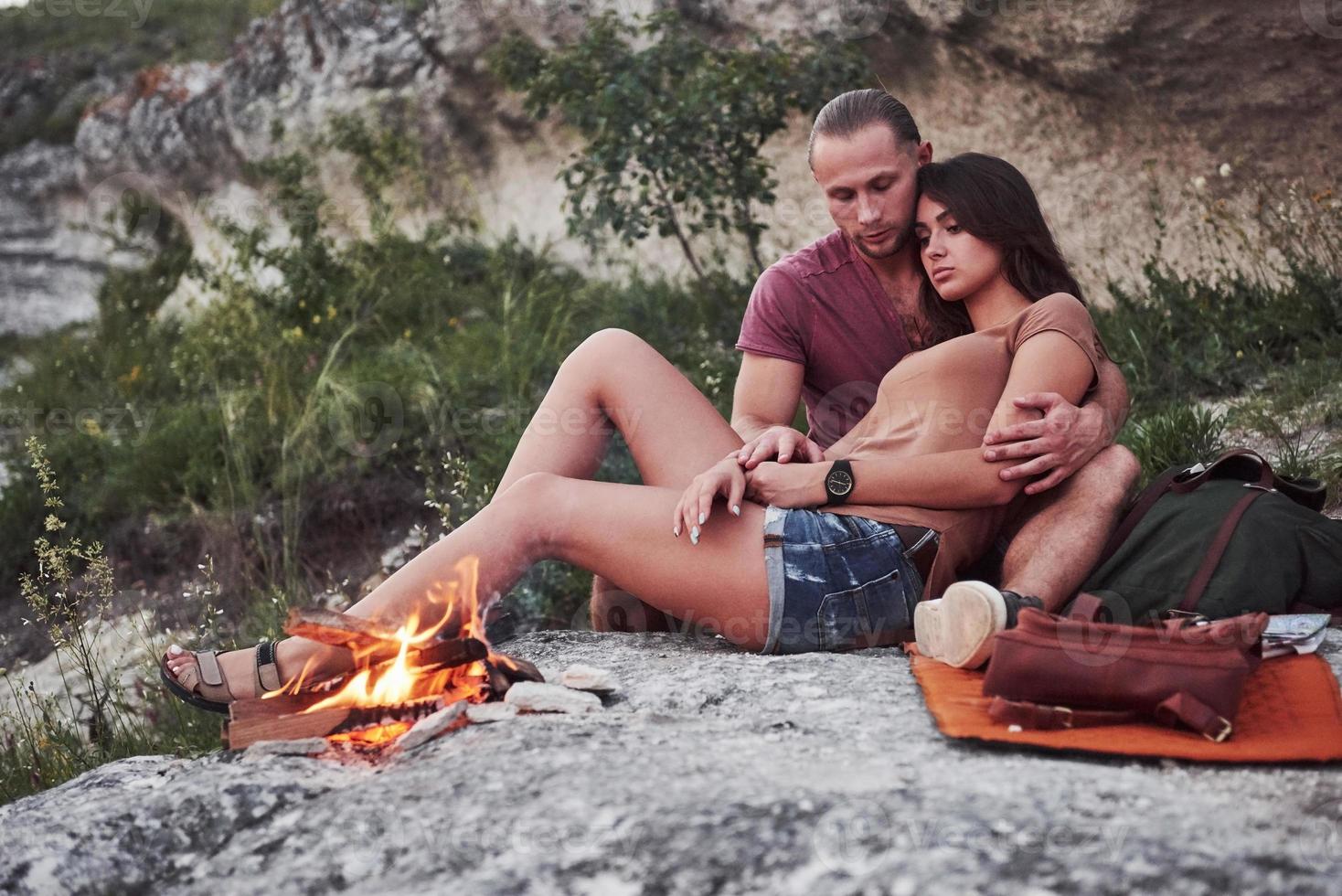 casal abraçando com mochila sentado perto do fogo no topo da montanha, apreciando a vista da costa de um rio ou lago. viajando ao longo das montanhas e da costa, conceito de liberdade e estilo de vida ativo foto